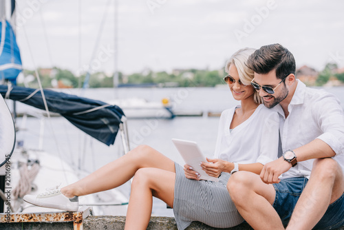Foto smiling young couple in sunglasses sitting and using digital tablet near yacht