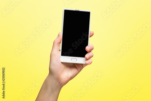 Mockup of female hand holding frameless cell phone with black screen isolated at yellow background.