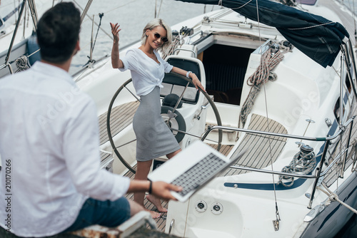 high angle view of smiling girl at steering wheel waving hand and looking at young man on yacht
