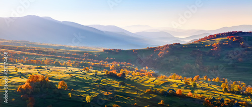 Fototapeta Naklejka Na Ścianę i Meble -  Extraordinary autumn landscape. Green fields with haystacks. Trees covered with orange and crimson leaves. Mountain landscapes.