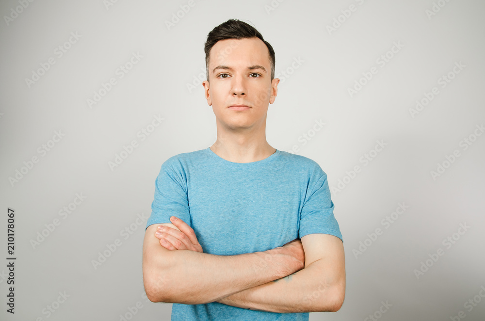Young guy with serious face and crossed arms dressed in a blue t-shirt stand on a light background. Young guy with serious face and crossed arms dressed in a blue t-shirt stand on a light background.
