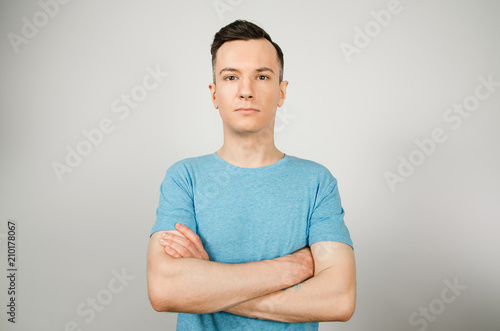 Young guy with serious face and crossed arms dressed in a blue t-shirt stand on a light background.