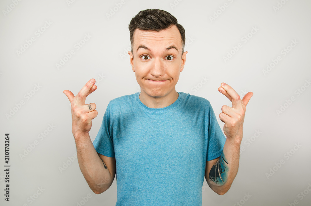 Young guy dressed in a blue t-shirt crossed fingers on a light background. Young guy dressed in a blue t-shirt crossed fingers on a light background.