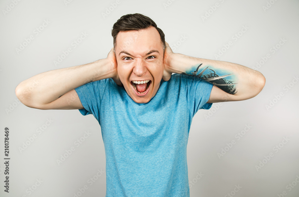 Young guy dressed in a blue t-shirt closing ears on a light background. Young guy dressed in a blue t-shirt closing ears on a light background.