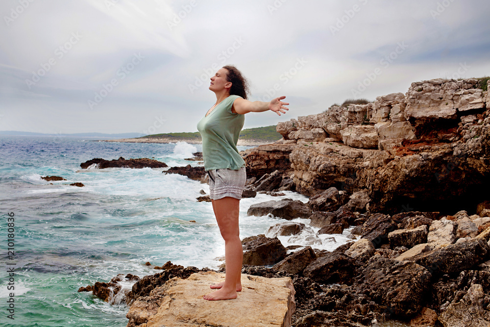 Woman with outstretched arms enjoying the wind and breathing fresh air on the rocky beach 