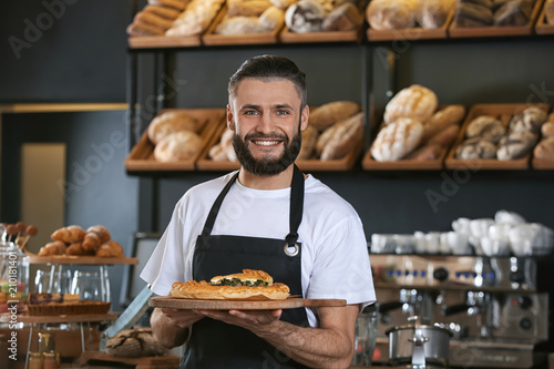 Male baker holding wooden board with delicious pie indoors