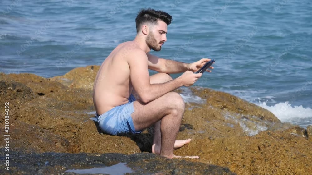 Young hansome shirtless man at sea reading with ebook reader or small tablet PC sitting on beach rocks Young hansome shirtless man at sea reading with ebook reader or small tablet PC sitting on beach rocks
