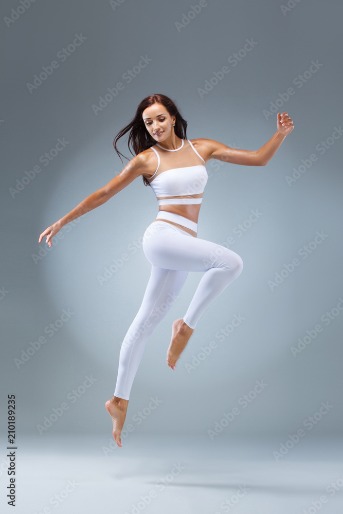 Fototapeta premium Attractive excited fitness girl, ballerina dancer in sportwear jumping of joy isolated over gray background.