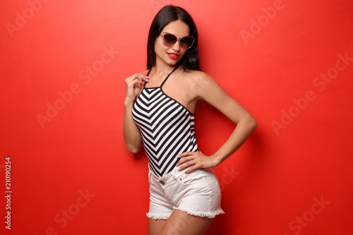 Beautiful young woman in swimming suit and shorts on color background