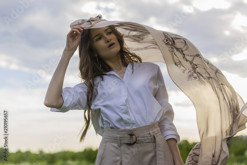 Beautiful stylish girl wearing a trendy white shirt, beige trousers and a pareo poses outdoors against a background of meadow full of wild flowers and blue cloudy sky