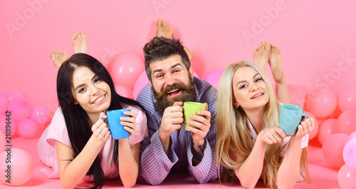 Threesome relax in morning with coffee. Lovers in bed concept. Lovers drinking coffee in bed. Man and women, friends on happy faces lay, pink background. Man and women in domestic clothes, pajamas.