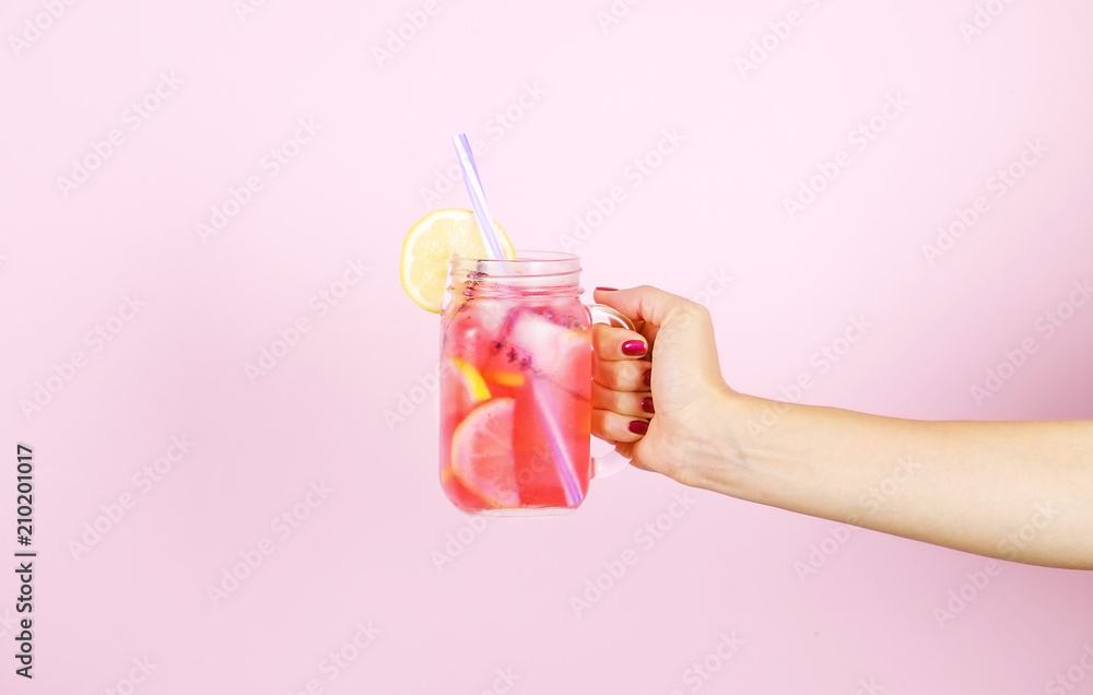 Young woman hands holding freshly squeezed strawberry lemonade of citrus fruits. Female with mason jar full of cold cocktail, lemon, orange, lime & mint leaves. Pink background, copy space, close up. - obrazy, fototapety, plakaty 