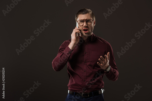 angry man in eyeglasses talking by smartphone isolated on black