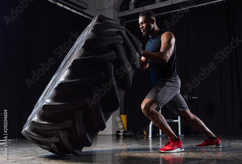 side view of athletic african american man exercising with tyre and looking away in gym