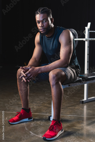 full length view of young african american sportsman sitting and looking at camera in gym