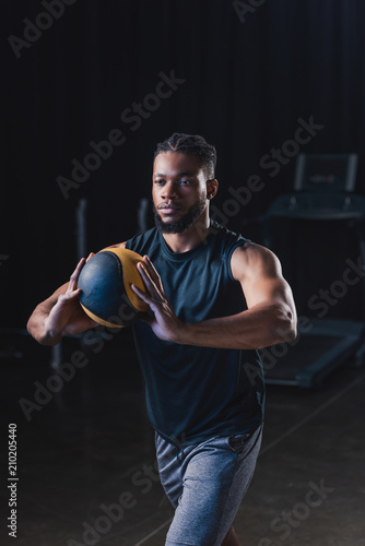 muscular african american sportsman holding ball while exercising in gym