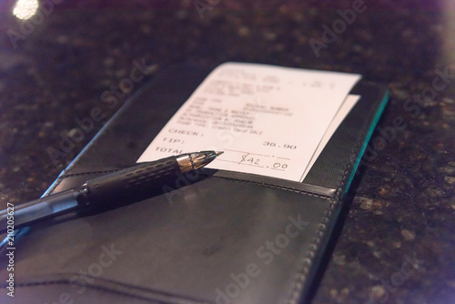 Open leather check holder with restaurant bill and pen. Top view, soft focus receipt with total amount on marble bar or pub table. Customer payment for dining service