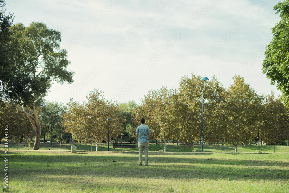 Minimalist portrait of man with his back to the distance in the middle of a landscape of a park or forest with many green trees and a large expanse of grass. Minimalist portrait of man with his back to the distance in the middle of a landscape of a park or forest with many green trees and a large expanse of grass.