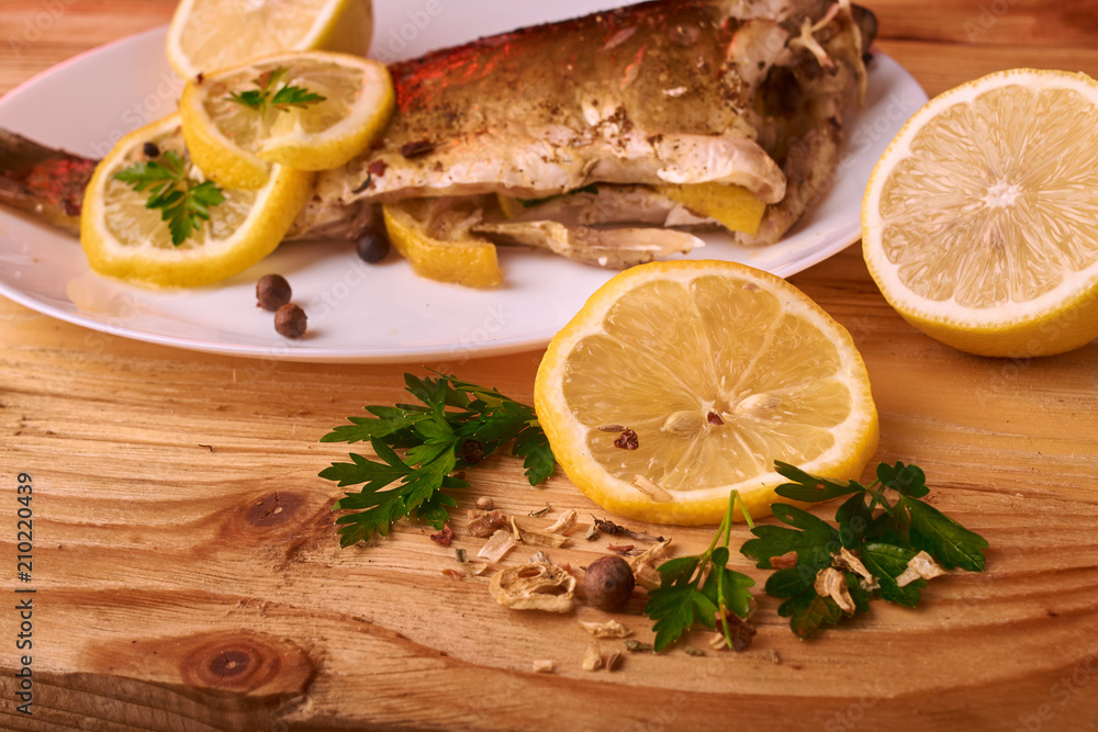 Baked fish with lemon, spices and herbs on white plate on wooden background without head Baked fish with lemon, spices and herbs on white plate on wooden background without head