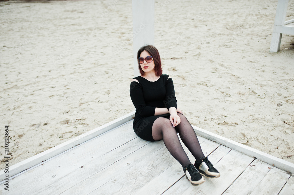 Portrait of brunette girl in black dress sitting at white wooden construction.