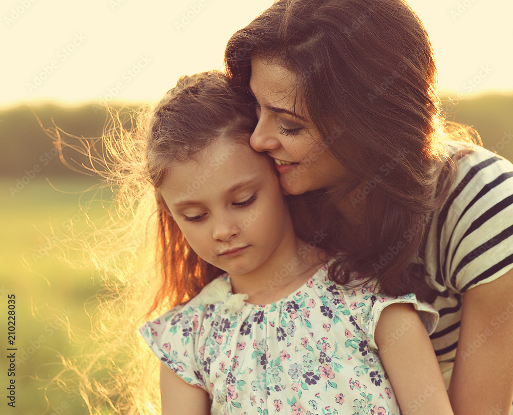 Emotional calm mother hugging her relaxing kid girl with loving eyes on sunset bright summer background. Closeup toned color portrait Emotional calm mother hugging her relaxing kid girl with loving eyes on sunset bright summer background. Closeup toned color portrait
