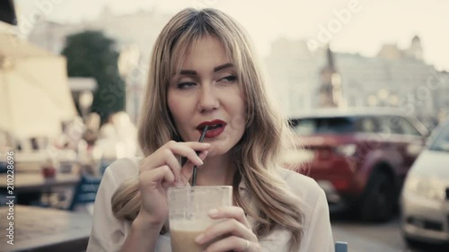 In Paris, a young Caucasian blonde woman, curly hair, with sexy red lips, drinks an alcoholic cocktail through a black straw from a large bar glass in a restaurant on a city street. The concept of