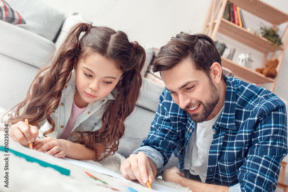 Father and little daughter at home lying on floor drawing together with color pencils concentrated Father and little daughter at home lying on floor drawing together with color pencils concentrated
