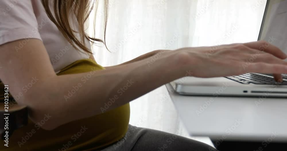 Crop side view of casual woman in expectancy sitting at working table and using laptop with smartphone Crop side view of casual woman in expectancy sitting at working table and using laptop with smartphone