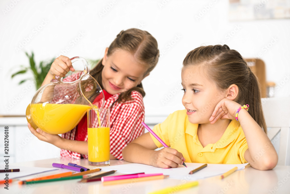 portrait of kid pouring juice into glass for sister at table with papers and pencils for drawing at home portrait of kid pouring juice into glass for sister at table with papers and pencils for drawing at home