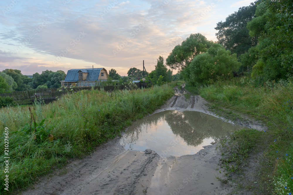 Vanishing dirt road with deep rut and puddles in village at sunset