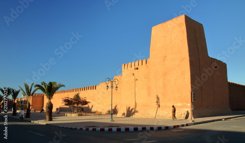Wysoki, poteżny mur marokańskim mieście, dekoracynie wykończony, z gliny pise, na ulicy kolkoro mieszkańców, lampy uliczne, palmy, intensywnie niebieskie niebo