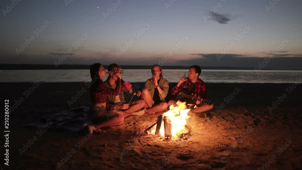 Accelerated footage of friends sitting on the beach on a plaid near the bonfire, clinking with beer bottles. Happy time, friendship. Night, wind blowing
