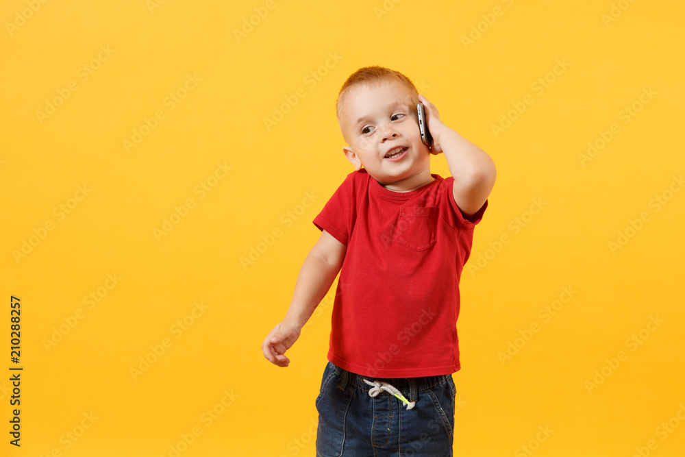 Little kid boy 3-4 years old in red t-shirt talking on mobile phone, conducting pleasant conversation isolated on yellow background. Kids childhood lifestyle concept. Problem of children and gadgets. Little kid boy 3-4 years old in red t-shirt talking on mobile phone, conducting pleasant conversation isolated on yellow background. Kids childhood lifestyle concept. Problem of children and gadgets.