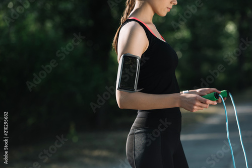 Cropped view of athletic woman with smartphone in armband holding jump rope in park photo