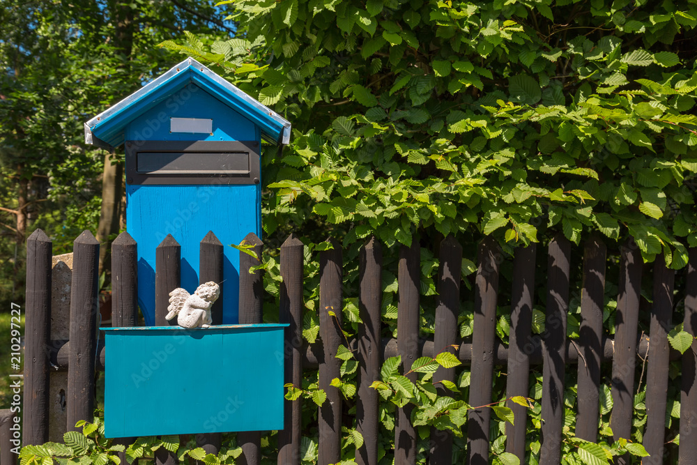 Blauer Briefkasten mit Engelsfigur an einem Zaun