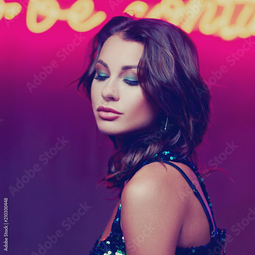 Fashion portrait of young beautiful woman with middle length dark hair wearing green sequins dress posing against red wall with neon letters in night bar
