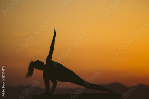 Fototapeta Naklejka Na Ścianę i Meble -  Young woman is practicing yoga on the mountain at sunset. Silhouette of young woman practicing yoga outdoor.