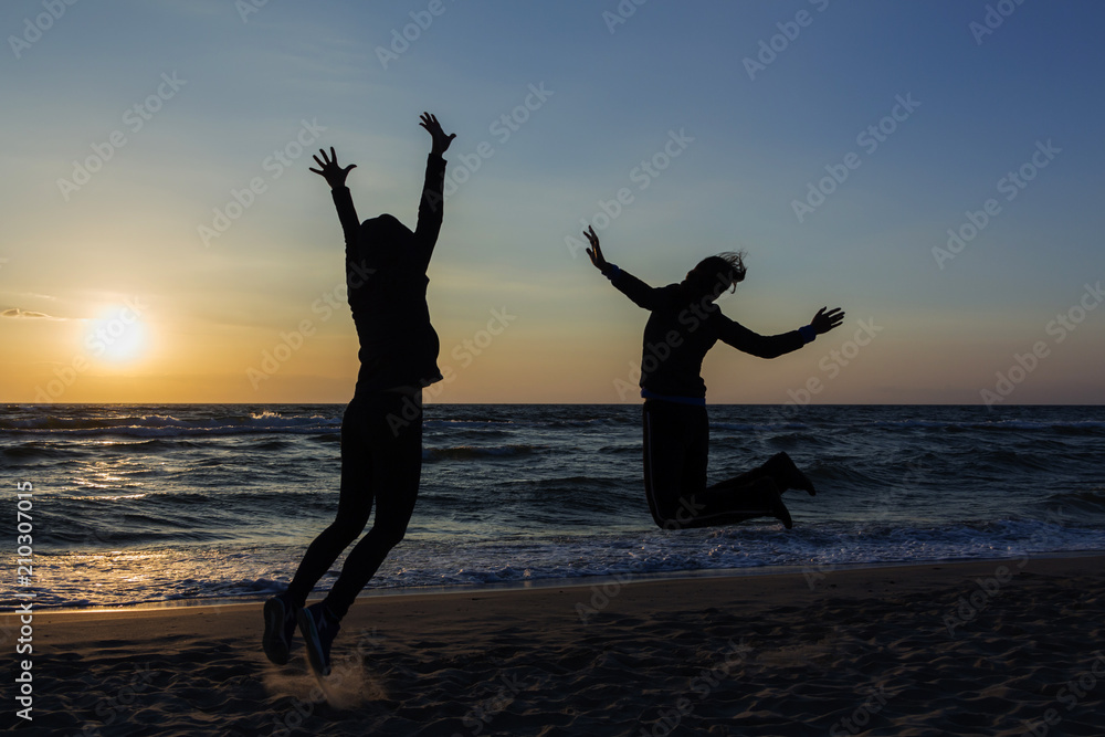 Fototapeta premium Two happy girls having fun and standing with raised arms on background of sunset sea.