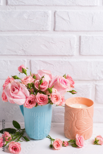 Pink roses flowers in blue cup  and burning candle against  white brick wall.