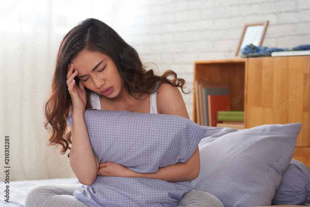 Portrait shot of beautiful Asian woman sitting on bed and holding pillow in hand while suffering from terrible headache, interior of cozy bedroom on background Portrait shot of beautiful Asian woman sitting on bed and holding pillow in hand while suffering from terrible headache, interior of cozy bedroom on background