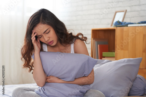 Portrait shot of beautiful Asian woman sitting on bed and holding pillow in hand while suffering from terrible headache, interior of cozy bedroom on background