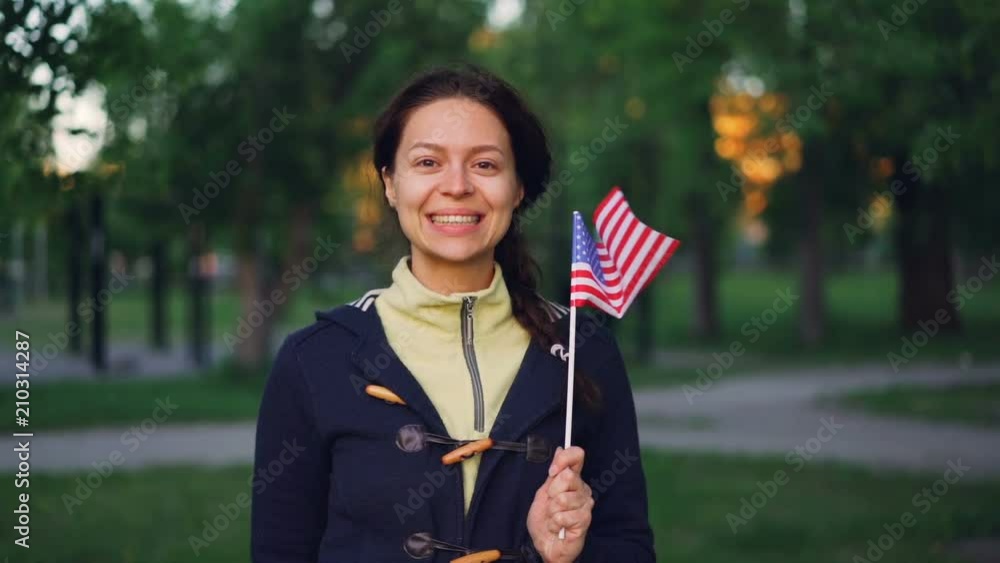 Slow motion portrait of pretty young lady American fan looking at camera and smiling waving flag of the United States. Happy people, countries and patriotism concept. Slow motion portrait of pretty young lady American fan looking at camera and smiling waving flag of the United States. Happy people, countries and patriotism concept.