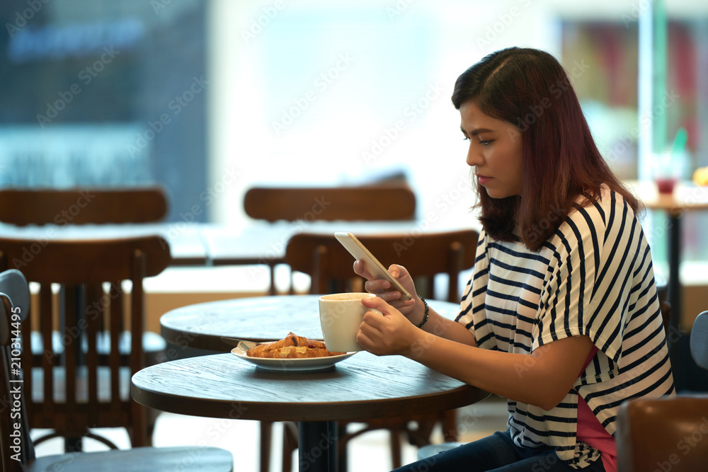 Profile view of beautiful Asian woman wearing striped T-shirt sitting at cafe table with cup of coffee and texting with friend on smartphone