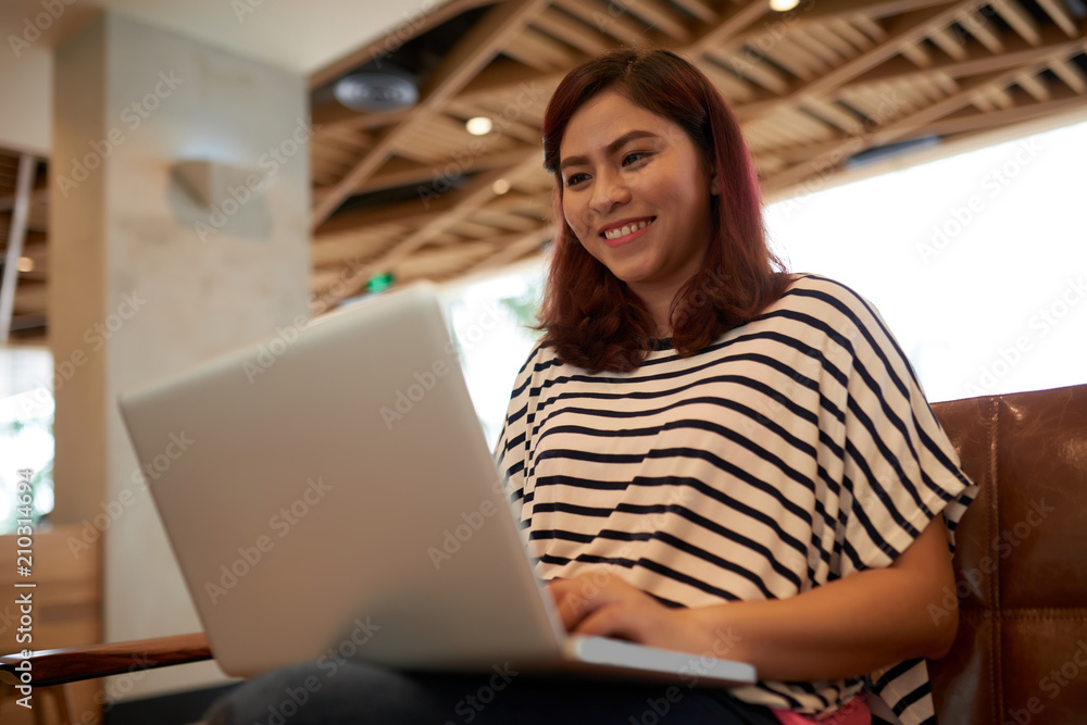 Cheerful Asian woman with charming smile sitting on cozy sofa and watching funny video on laptop, portrait shot Cheerful Asian woman with charming smile sitting on cozy sofa and watching funny video on laptop, portrait shot