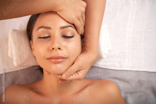 Young pleased woman enjoying facial massage with calming effect in beauty salon