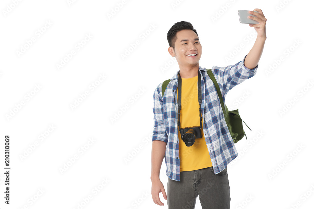 Cheerful Asian tourist with backpack taking selfie on modern smartphone while standing against white background, copy space Cheerful Asian tourist with backpack taking selfie on modern smartphone while standing against white background, copy space
