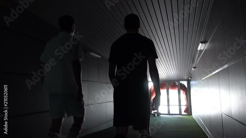Two football players are walking along a dark tunnel to the football field. View from the back
