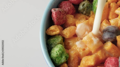 Top close view of adding milk to a large blue bowl of dry fruit flavor breakfast cereal on a white counter top. 