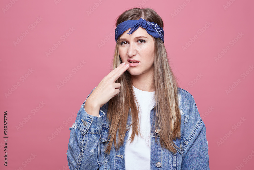 Beautiful hippie lady poses against pink wall, makes sick and tired gesture, has displeased expression, wears stylish headband, denim oversized jacket. Negative human emotions, body language concept. Beautiful hippie lady poses against pink wall, makes sick and tired gesture, has displeased expression, wears stylish headband, denim oversized jacket. Negative human emotions, body language concept.