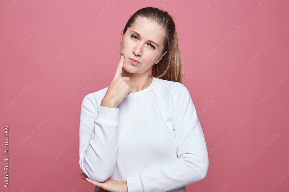 Indoor shot of attractive pensive female with long hair, looks foxy at camera, thinks about something, has pleasant appearance, isolated over pink background with copy space for promotional text. Indoor shot of attractive pensive female with long hair, looks foxy at camera, thinks about something, has pleasant appearance, isolated over pink background with copy space for promotional text.
